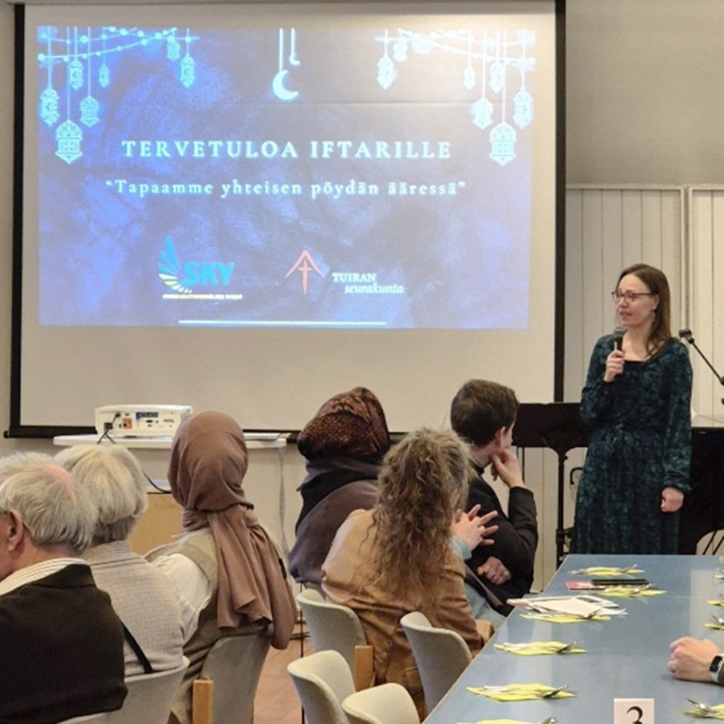 Nainen puhuu yleisölle, kankaalle heijastetussa diassa toivotetaan tervetulleeksi tapahtumaan.